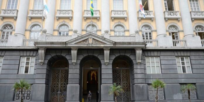 Facade of a historic building with tall columns, arched doorways, and ornate iron gates; flags on the balcony above the entrance.