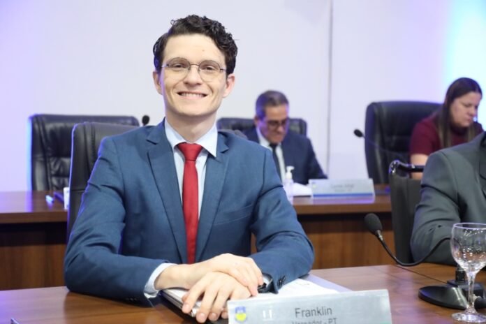 Smiling man in a blue suit and red tie sits at a conference table; nameplate reads 'Franklin'.