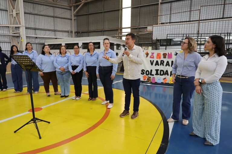 Man with microphone addressing a line of women in light blue shirts inside a gymnasium; a colorful banner behind them reads something like 'VARI... SOLIDÁR' and a music stand is visible to the front left.