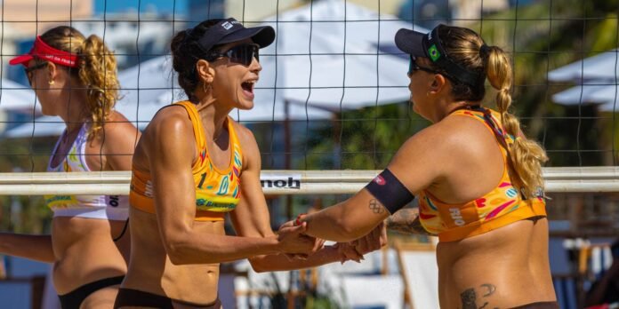 Two female beach volleyball players in orange uniforms shake hands at the net after a point on a sunny court.