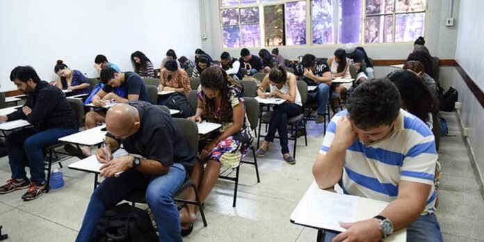 Students seated in a classroom taking a written exam, heads bent over their desks.