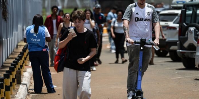 Man on a blue electric scooter rides along a busy sidewalk as pedestrians walk behind him in an urban area.