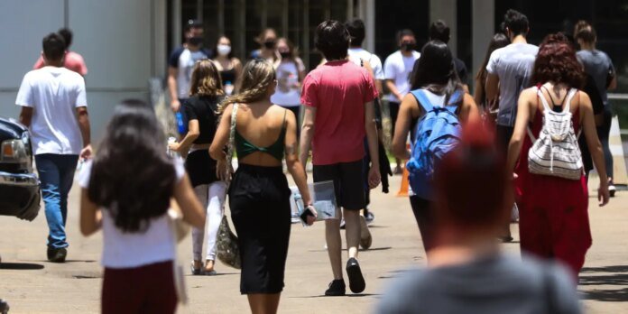 People walk away from the camera in a sunny outdoor campus setting, carrying backpacks and bags.