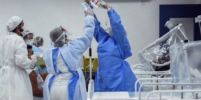 Healthcare workers in full PPE (gowns, masks, face shields) gather around a patient bed in a hospital ICU. They manage IV lines and equipment.