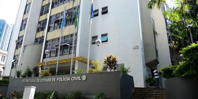 Facade of a government building with multiple flags and a sign reading 'SECRETARIA DE ESTADO DE POLICIA CIVIL'; a man stands on stairs to the right, taking a photo.