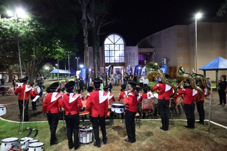 Marching band in red jackets with white accents performs at night outdoors, drums and tubas visible, with spectators and blue tents in the background.