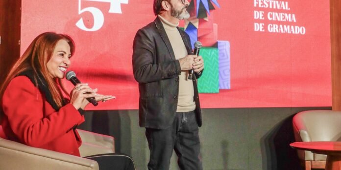 Two presenters on stage at a cinema festival; woman in a red coat smiles and speaks into a microphone while a man in a beige turtleneck and blazer stands beside her against a red backdrop.