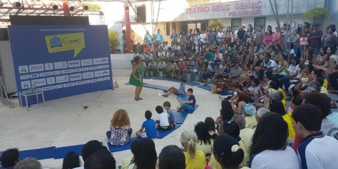 Audience gathered around a circular stage as a woman in a green dress addresses children seated on blue mats; sponsor backdrop on the left.