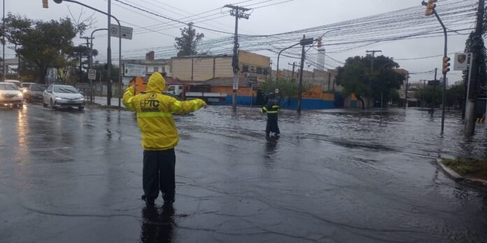Two people in rain gear standing in a flooded street directing traffic during a heavy rain. A yellow jacket labeled EPIC is visible in the foreground, with cars and flooded storefronts in the background.