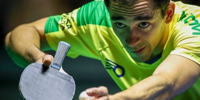 Close-up of a man in a neon yellow shirt intensely focused, reaching with a table tennis paddle to strike a ball.
