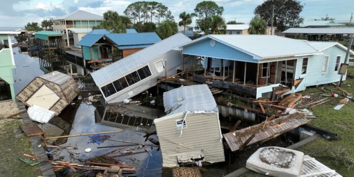 Aerial view of a flooded neighborhood with collapsed and tilted houses, scattered debris, and standing water after a disaster.