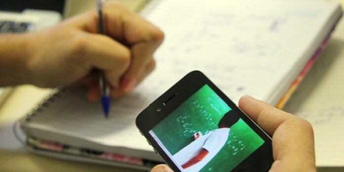 Close-up of a smartphone screen showing a teacher at a chalkboard, while a person writes in a notebook in the background, capturing a learning scene.