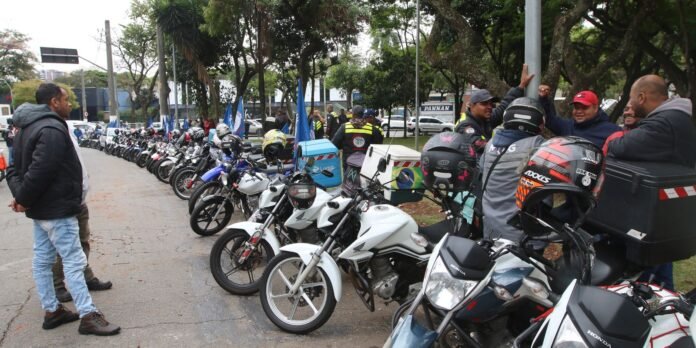 Row of parked motorcycles along a street, with riders standing nearby and chatting near a park.