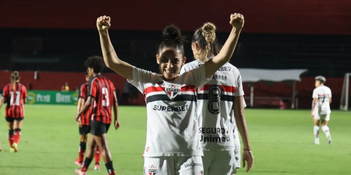 Female soccer player in a white jersey with red and black stripes raises her arms in celebration on a green pitch, teammates blurred in the background.