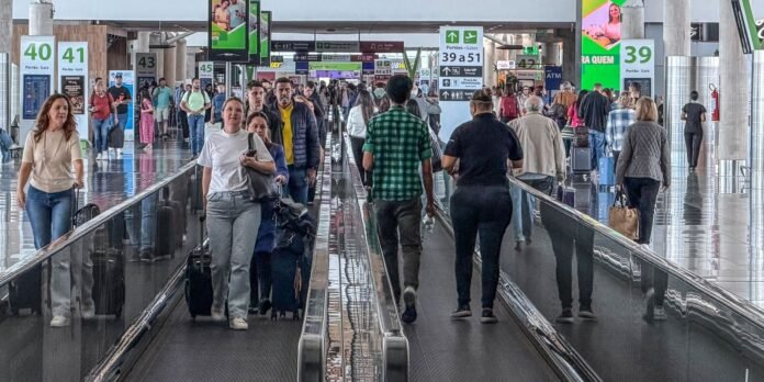 Crowded airport terminal with travelers on a moving walkway carrying luggage and backpacks.