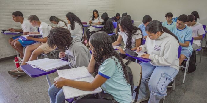 Group of diverse students seated at purple desks in a classroom, focused on notebooks and worksheets while taking notes.