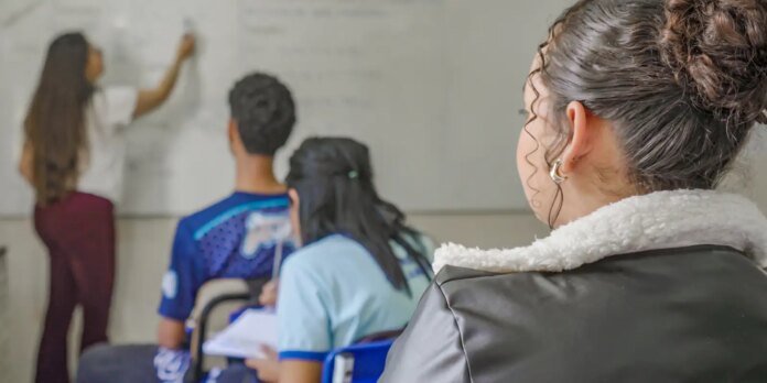 Student with a curly bun and silver hoop earrings sits in a classroom, facing a teacher at the whiteboard in the background.
