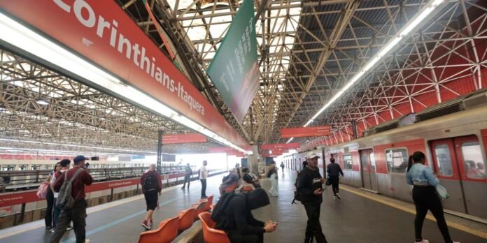 Platform at Corinthians-Itaquera station with commuters waiting as a silver train sits on the right under a red sign. 1/2? (informative)