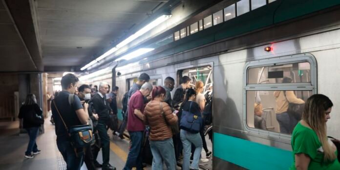People board a silver subway train with a teal stripe at a busy underground station; some passengers wear masks and carry bags.