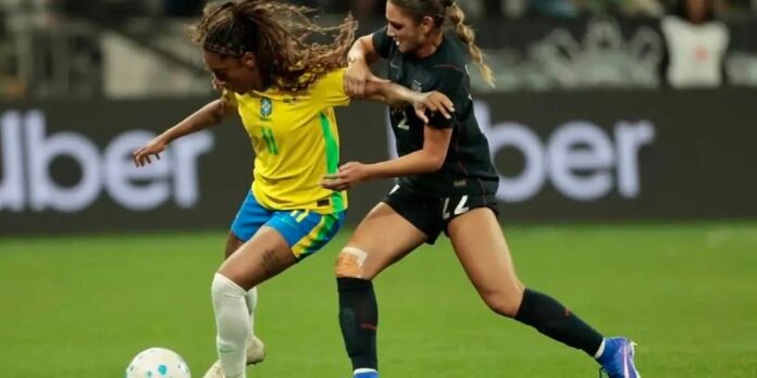 Two female soccer players chase the ball during a match; one in a yellow Brazil jersey and blue shorts, the other in a black kit, tussling for possession.