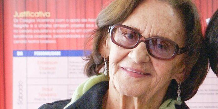 Smiling elderly woman with short hair and pink glasses, wearing earrings and a green scarf, indoors with a red poster in the background.