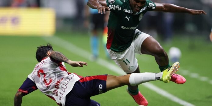 Soccer players in a slide tackle during a match, one in white/red, the other in green, on a grassy field.