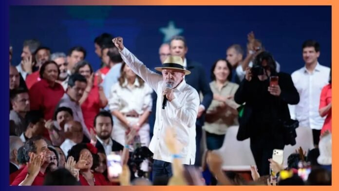 Man in a white shirt and straw hat speaks into a microphone on stage, fist raised, surrounded by a cheering crowd at a rally.