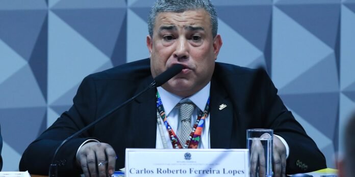 Panelist in a dark suit speaks into a microphone at a conference, with a nameplate and water glass in view.