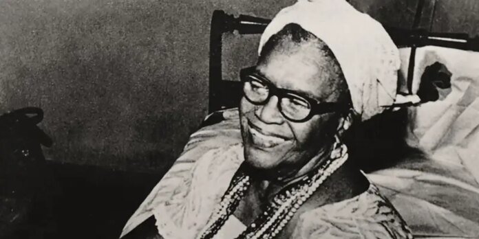 Elderly woman with thick glasses and a white headscarf sits in a hospital bed, smiling, wearing multiple bead necklaces.