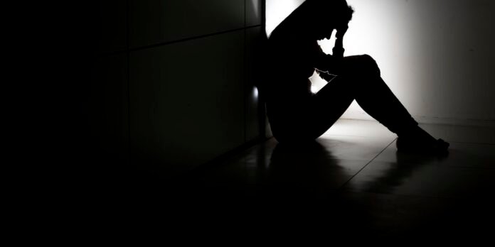 Silhouette of a person sitting on the floor by a wall, head in hands, conveying distress or despair in a dark setting.