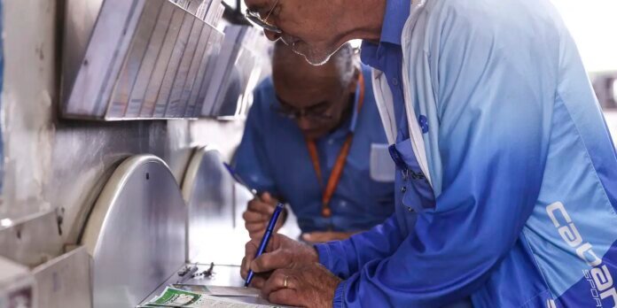 Two older men in blue work uniforms sign a document on a metal machine console.