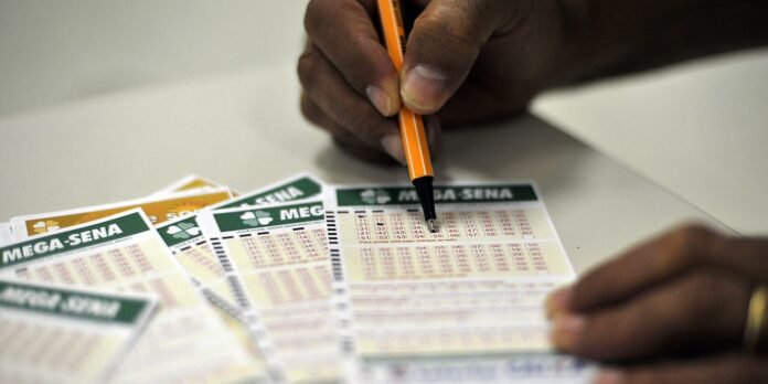 Person's hand writes on MEGA-SENA lottery tickets with an orange pen at a desk.