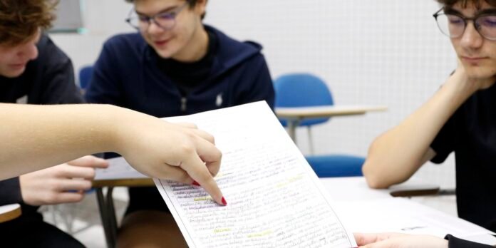 Close-up of a hand with red nails pointing to a handwritten, highlighted worksheet while two students read in a classroom.
