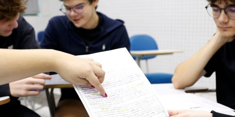 Close-up of a hand with red nails pointing to a handwritten, highlighted worksheet while two students read in a classroom.