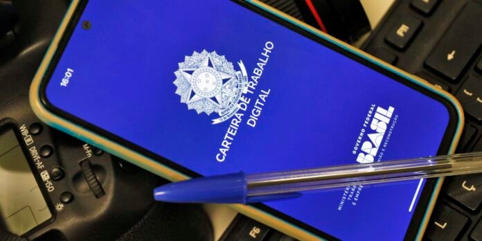 Smartphone screen shows the Brazilian 'Carteira de Trabalho Digital' logo on a blue background, resting on a laptop keyboard with a blue pen across it.