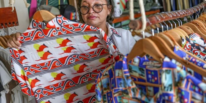Woman with round glasses holds up a white shirt with red and yellow patterns in a clothing stall at a market.