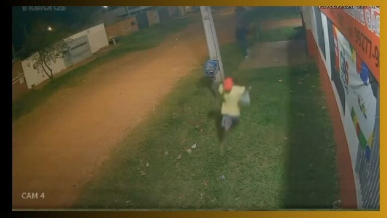 Nighttime security footage of a person wearing a red helmet and light-colored jacket riding a small motorbike along a grassy area beside a wall with posters; blurred motion suggests movement toward camera.