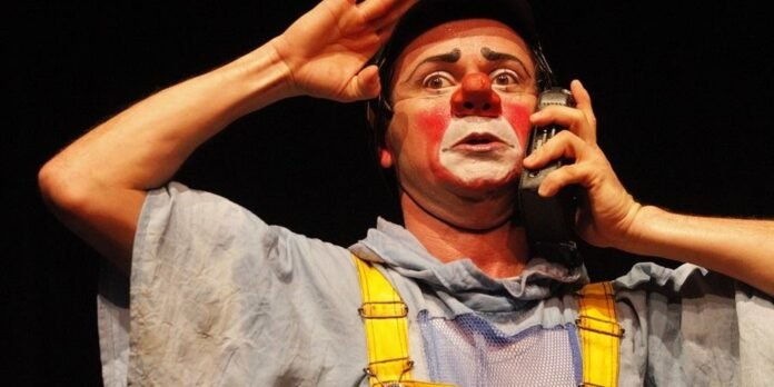 Clown with red nose and white face paint holds a cellphone to his ear on stage, wearing yellow suspenders and a light shirt.
