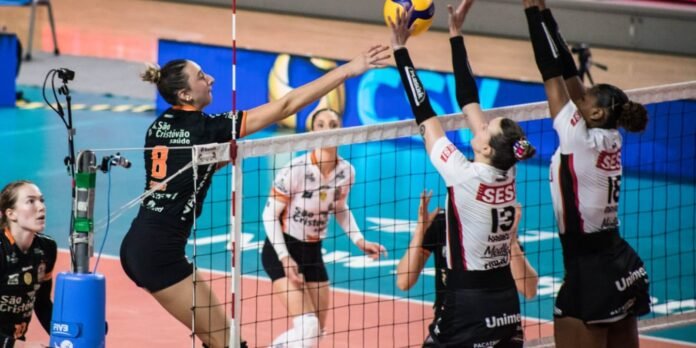 Female volleyball players at the net during a match; a black-uniform spiker (#8) leaps to hit the ball while white-uniform blockers jump to block the shot.