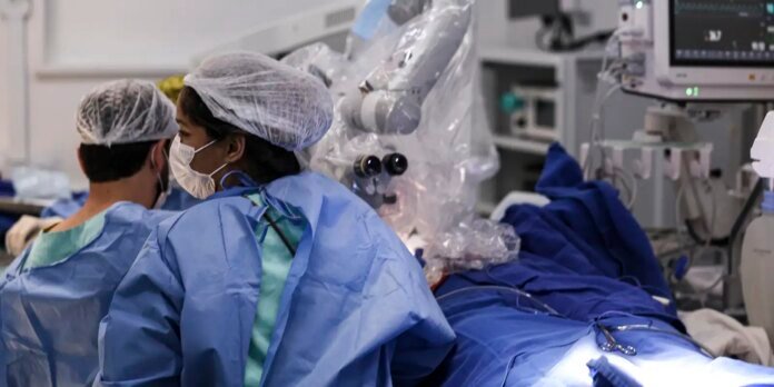 Surgeons in blue gowns and masks operate in a well-equipped operating room using a surgical microscope.