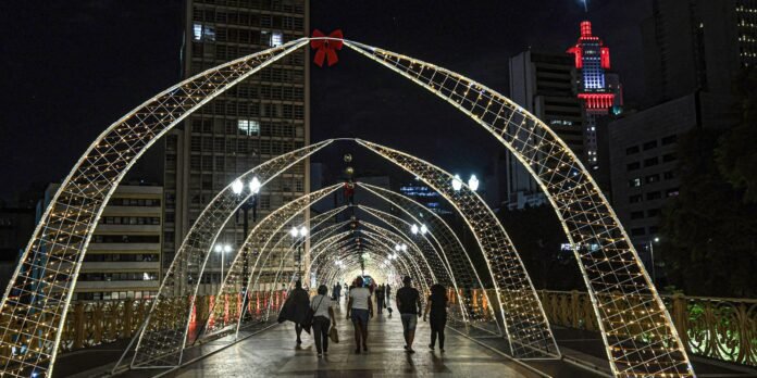 Night view of a pedestrian bridge with illuminated lattice arches forming a tunnel, people walking toward the city center, tall buildings in background with red-lit top.