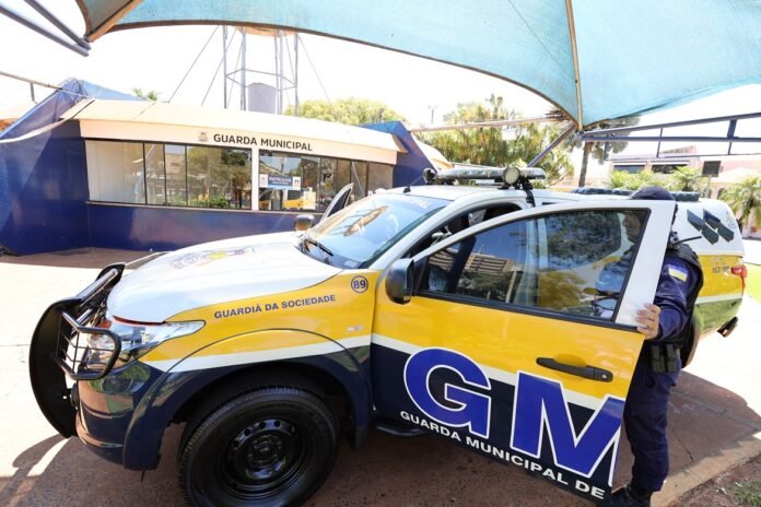 Municipal guard SUV with large GM logo parked near a guard building, officer opening the front door under a blue shade structure.