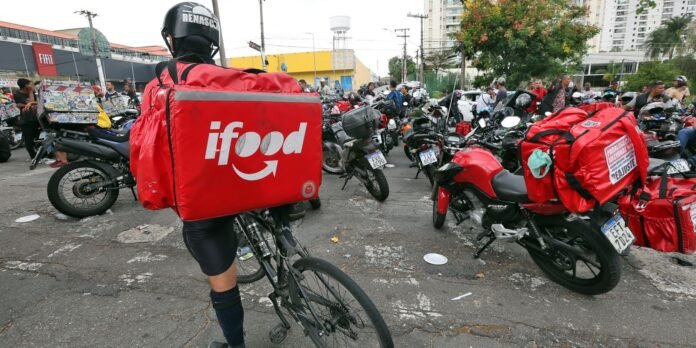 Delivery rider wearing a helmet with a large red iFood bag on their back pedals through a crowded street full of motorcycles and onlookers.