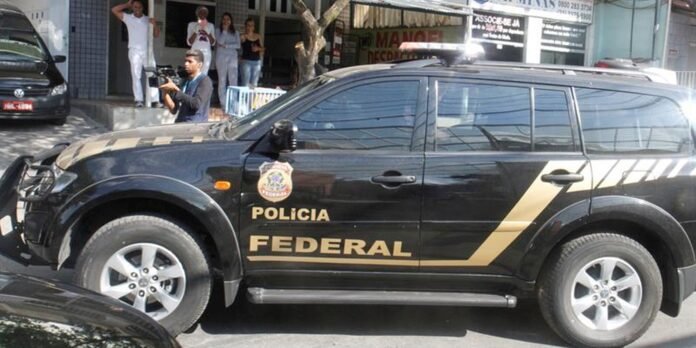Black Polícia Federal SUV with gold stripe parked on a street; a cameraman and onlookers stand in the background.