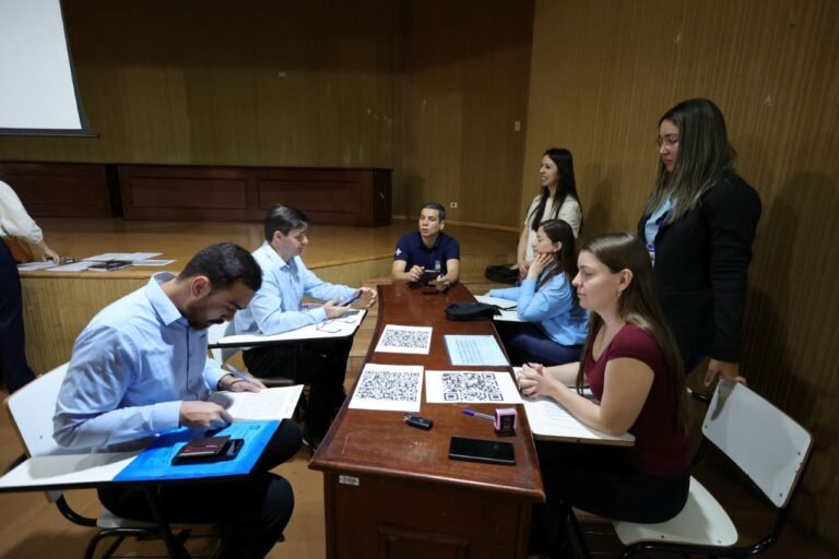 Group of six adults seated around a wooden table with documents and QR codes, in a conference room; two people stand nearby.