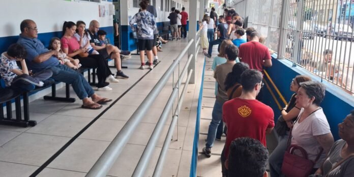Crowded transit terminal corridor with people seated along a blue wall on the left and others standing by a railing on the right, waiting to board or be called.