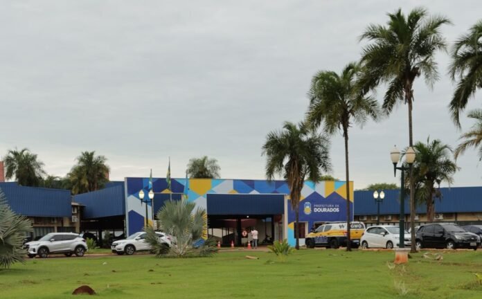 City hall in Dourados with a blue geometric mural, parked cars, and palm trees outside nonchalantly under a cloudy sky.