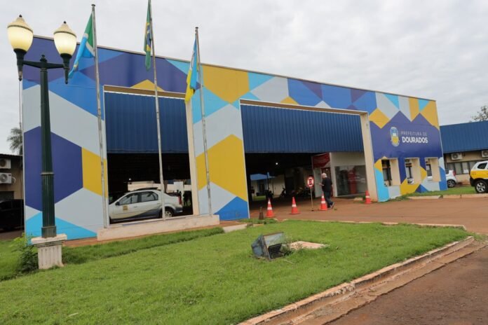 Municipal building with colorful geometric mural, flags, and open bays (Dourados City Hall) storefront area with cones and people nearby