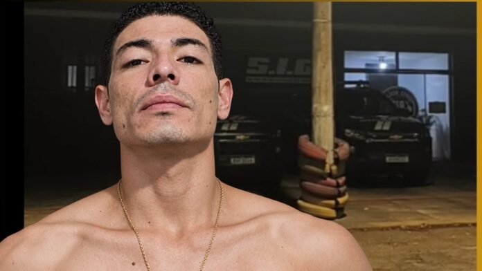 Close-up portrait of a shirtless man with a gold chain, looking slightly upward, in a gym with cars in the background.