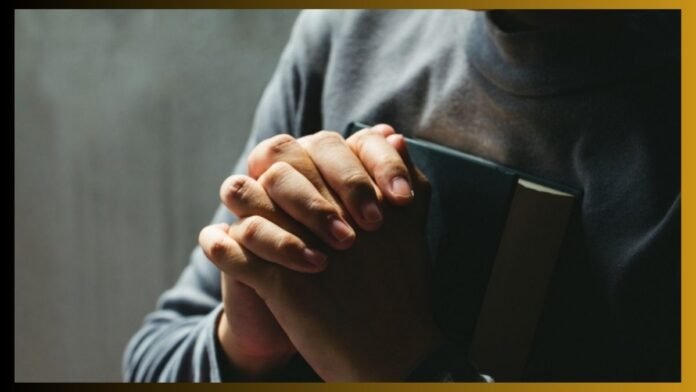 Person’s hands pressed together in prayer while clutching a closed book against a dark background.